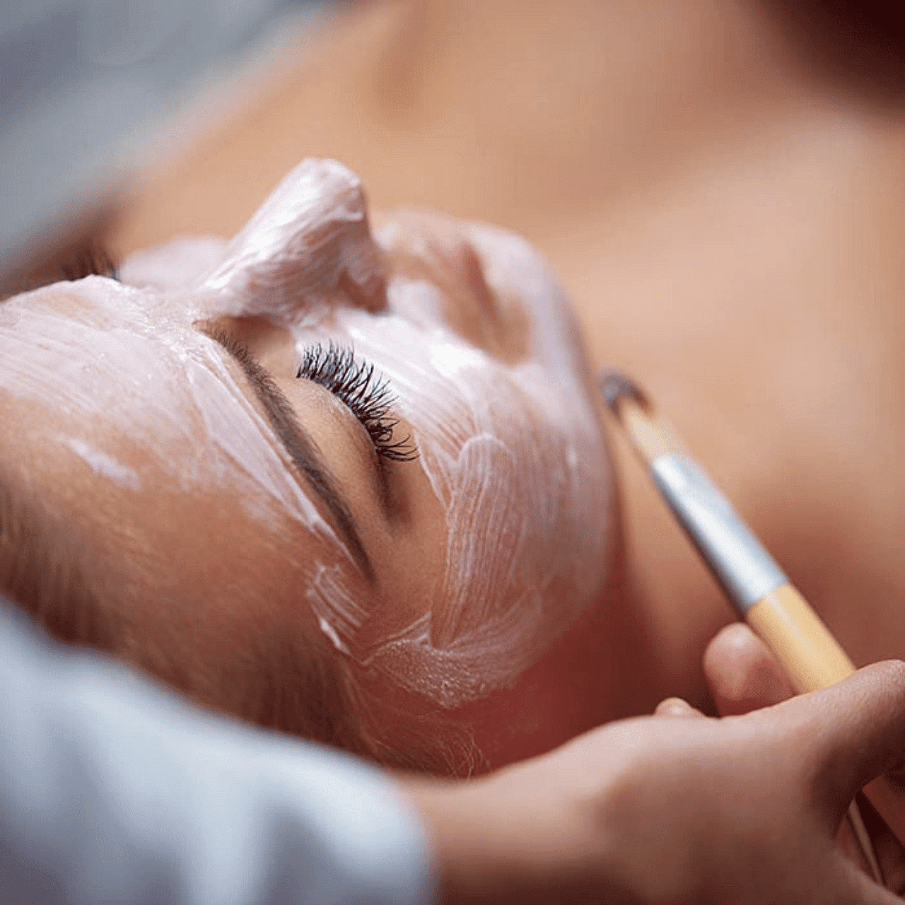 Person receiving a facial treatment with a brush applying a white mask on their face.