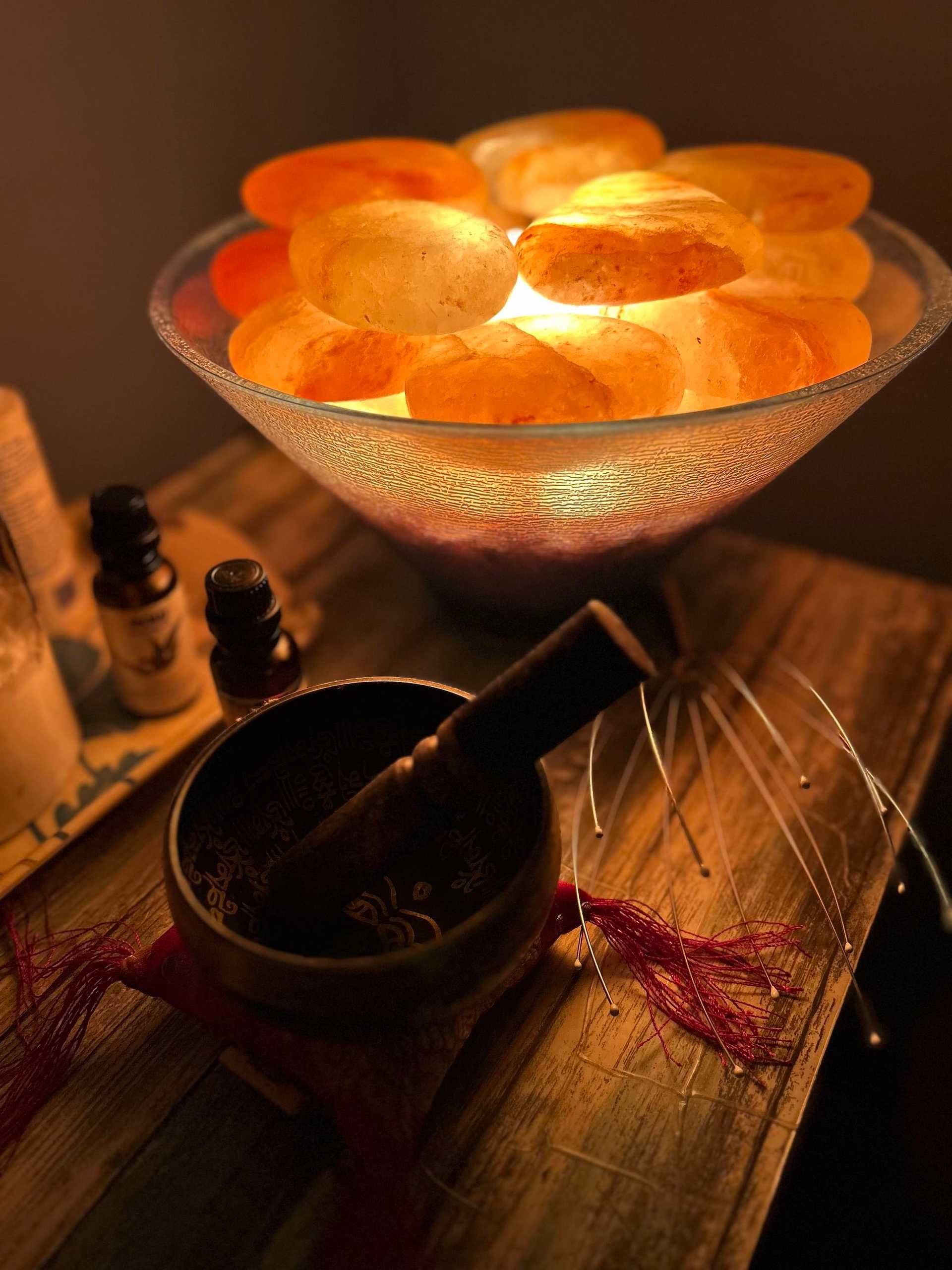 Lit salt lamp with Himalayan salt stones beside essential oils and a Tibetan singing bowl.